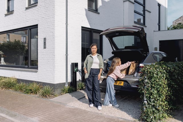 Vrouw staat voor huis bij elektrische auto en laadpaal 